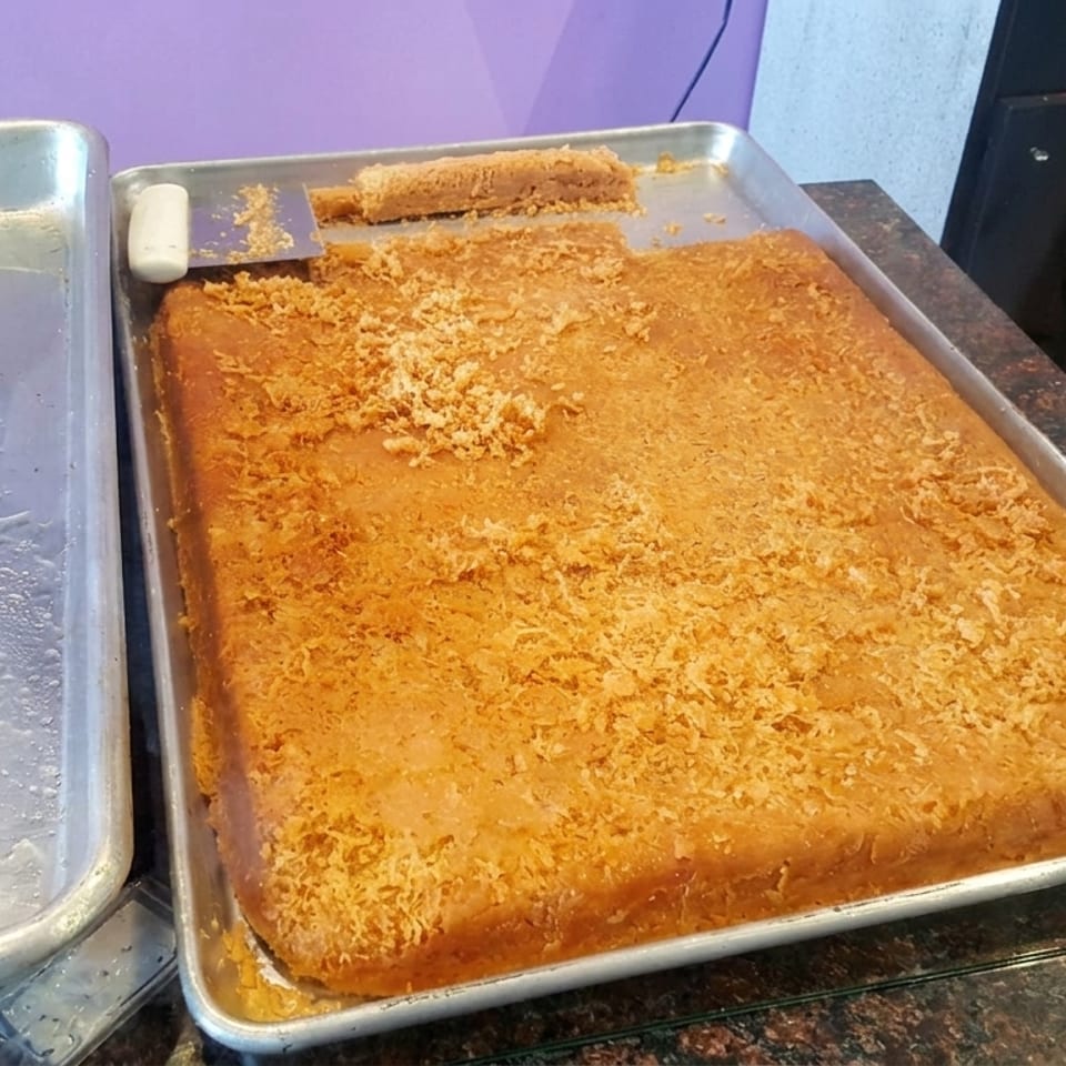 Large golden rectangular baked pastry with a crumbly topping sits in a metal serving tray at a bakery counter.