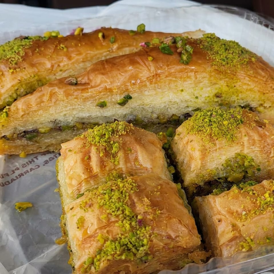 Assorted golden baklava pastries topped with crushed pistachios in a clear plastic container.